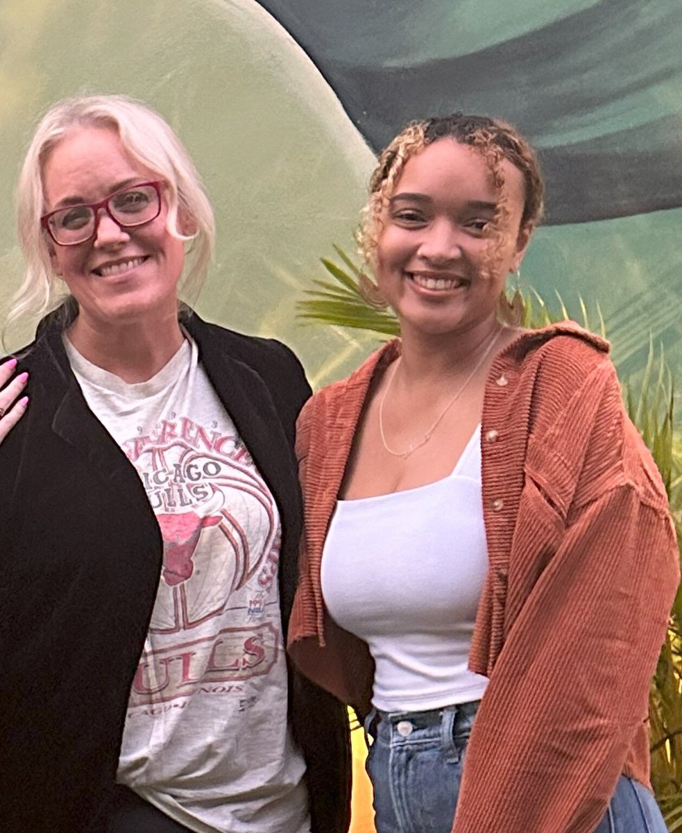 Co-founders Dana and Abby in San Francisco, standing in front of wall mural painted with lush tropical leaves; cropped image shows from the waist up