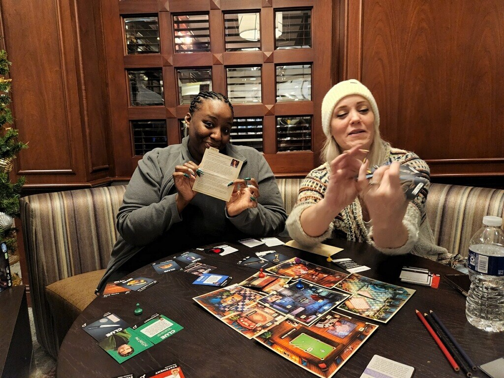 Co-founders Kernika and Dana sitting at a small table in a hotel lounge, playing a board game. Kernika is holding a clue card in her hand and dana is looking at an object she's holding.